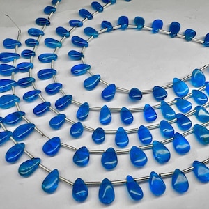 Könnte beinhalten: Stränge aus blauen, tropfenförmigen Perlen. Die Perlen sind auf silberfarbenem Draht aufgefädelt. Die Perlen haben eine glänzende Oberfläche und variieren leicht in der Schattierung, von hellblau bis tiefblau. Die Perlen sind in Schichten angeordnet.