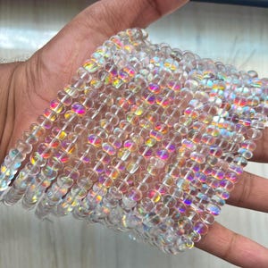 Könnte beinhalten: Stränge aus klaren, irisierenden Glasperlen. Jede Perle ist rund und hat einen Durchmesser von etwa 0,6 cm. Die Perlen reflektieren einen Regenbogen aus Farben, darunter Rosa, Gelb und Blau. Die Perlen sind aufgefädelt und erzeugen einen schimmernden Effekt.
