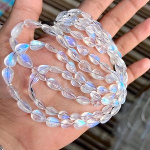 Può includere: Una collana di perline iridescenti a forma di goccia. Le perline trasparenti hanno una sottile lucentezza arcobaleno, infilate per formare una collana o un braccialetto. Le perline sono tenute in una mano, mostrando il loro colore e la loro forma.