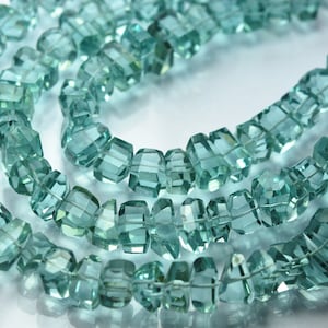 Puede incluir: Una hebra de cuentas de piedras preciosas facetadas de color azul claro. Las cuentas están enhebradas en un hilo transparente y están dispuestas en un patrón suelto y fluido.