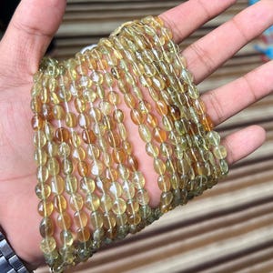 以下が含まれることがあります： 楕円形の黄褐色宝石ビーズのコレクション。ビーズはつながっており、複数のストランドを形成しています。宝石は半透明で、薄い黄色から濃い茶色まで色合いが異なります。ビーズは手で持たれています。