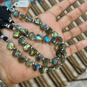 May include: Three strands of faceted labradorite gemstone beads. The beads display a range of colors, including green, blue, and gold, with a metallic sheen. Each strand is finished with a black tassel and silver accents, creating a unique jewelry piece.