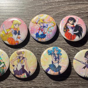 Op de afbeelding: Tien ronde buttons met kleurrijke illustraties in anime-stijl van verschillende personages in sailor-outfits. De buttons hebben een glanzende afwerking en zijn gerangschikt op een houten oppervlak.
