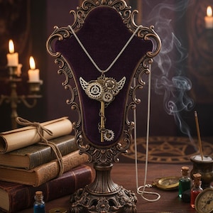 Può includere: Una collana con pendente a chiave in stile steampunk con intricati dettagli di ingranaggi e ali, esposta su un supporto decorativo con retro in velluto viola. Il supporto è ornato, con finitura bronzo. La scena comprende candele, libri e altri accessori vintage.