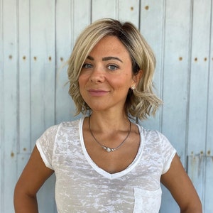 May include: A woman wearing a white, semi-sheer V-neck t-shirt with a pocket. She has short, wavy blonde hair and is wearing a silver necklace with three small, round pendants. A pink bracelet is visible on her wrist.