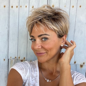 Op de afbeelding: Een vrouw met kort, blond gelaagd haar en blauwe ogen glimlacht, draagt een witte V-hals T-shirt en een zilveren ketting met drie bloemvormige hangers. Ze raakt haar oor aan en een kleine, ronde oorbel is zichtbaar.