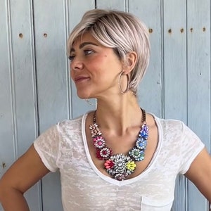 Könnte beinhalten: Ein auffälliges Statement-Collier mit großen, blütenförmigen Anhängern in Rosa, Rot, Blau, Gelb und Weiß. Das Collier wird mit einem weißen T-Shirt und silbernen Creolen getragen. Der Hintergrund ist eine hellblaue Holzwand.