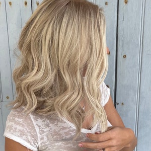 Peut inclure: Cheveux blonds mi-longs avec une texture ondulée. Les cheveux ont des reflets et des mèches, créant de la dimension. La personne porte un t-shirt blanc semi-transparent. L'arrière-plan est un mur en bois bleu clair.