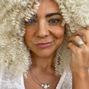 May include: A silver necklace with three clear, round, sparkling stones. The necklace is worn with a matching ring. The person in the image has blonde curly hair and is wearing a white top.