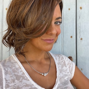May include: A silver necklace with three round, clear gemstones. The necklace is worn over a white, v-neck t-shirt. The person in the image has brown hair and is smiling.