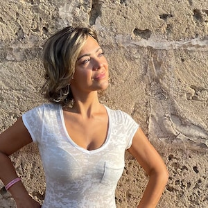 May include: A woman wearing a white, semi-sheer V-neck t-shirt with a small pocket. She is standing in front of a textured, light brown stone wall. She has short, light brown hair and is wearing a pink bracelet.
