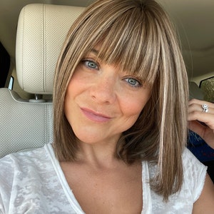May include: A woman with light skin and blue eyes, wearing a white V-neck shirt, smiles at the camera. Her short hair is a mix of blonde and brown with bangs. She is sitting in a car.