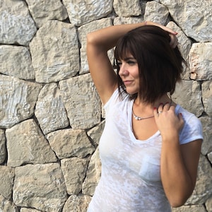 May include: A woman wearing a white V-neck t-shirt and a silver necklace with small, decorative elements. She is leaning against a stone wall, with her arm raised, and her dark hair is styled in a bob.