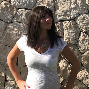 May include: A white V-neck t-shirt with a chest pocket. The shirt has a subtle, textured pattern. A silver necklace with three small, round pendants is visible. The background is a stone wall.