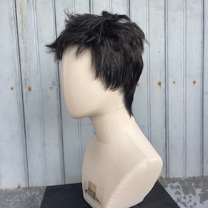 Könnte beinhalten: Dunkelbraune Perücke mit kurzem, gestuftem Stil, präsentiert auf einem Schaufensterkopf. Die Perücke hat ein natürliches Aussehen mit strukturierten Strähnen. Die Schaufensterpuppe ist cremefarben und vor einem verwitterten, hellblauen Holzgrund platziert.