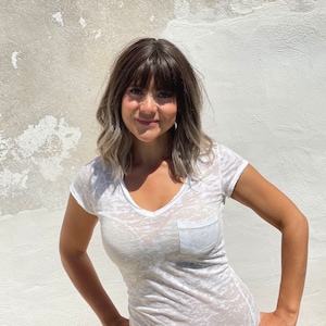 May include: A woman wearing a white, sheer V-neck t-shirt with a chest pocket. The woman has brown hair with blonde highlights and bangs. She is standing in front of a textured, off-white wall.
