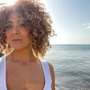 May include: A woman with curly blonde hair and a white tank top stands near the ocean on a sunny day. The image is a close-up, with the sea and sky visible in the background. The sun shines brightly.