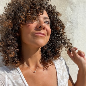 May include: A person with curly brown hair and a silver necklace with a small, round pendant. The person is wearing a white top. The background is a textured, off-white wall.
