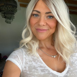 Op de afbeelding: Een persoon met lichtblond haar en blauwe ogen draagt een witte V-hals T-shirt en een zilveren ketting. Het haar heeft donkere wortels en is gestyled in losse golven. De persoon glimlacht. Op de achtergrond is een kroonluchter te zien.