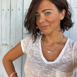 May include: A woman wearing a white V-neck t-shirt and a silver necklace with three circular pendants. She has short, wavy brown hair and is wearing two bracelets, one pink with the text "MASTOCITOSIS" and another with the text "VIVA VIVA".