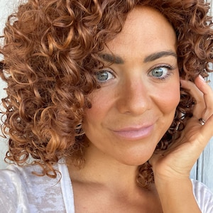 May include: A close-up portrait of a person with light skin, blue-gray eyes, and a warm complexion. The person has curly, reddish-brown hair. They are wearing a white top and gently touching their face with their hand.