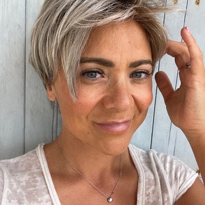 Könnte beinhalten: Eine Frau mit kurzen, gestuften, aschblonden Haaren und blauen Augen. Sie trägt ein weißes, strukturiertes T-Shirt und eine zarte Silberkette mit einem kleinen, runden Anhänger. Der Hintergrund ist eine hellblaue Holzwand.