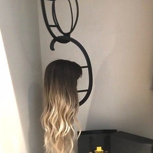 May include: A black metal wig stand holding a wig with dark roots and blonde highlights. The wig is in a white closet. Two black boxes and a black shopping bag with gold text are on the shelf below.