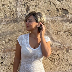 May include: A white, semi-sheer t-shirt with a V-neck and a pocket. The shirt has a subtle, textured pattern. The person is wearing a blonde wig with short, layered hair. The background is a textured, light brown wall.