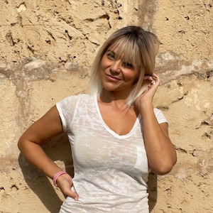 May include: A woman wearing a white, sheer V-neck t-shirt and a pink bracelet. She is standing in front of a textured, light brown wall. Her hair is blonde with bangs.