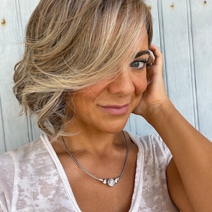 May include: A woman with blonde and brown layered hair, wearing a white V-neck shirt and a silver necklace with a central circular pendant and two smaller beads. The background is a light blue wooden wall.