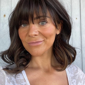 May include: A woman with dark brown hair and bangs, styled in loose waves. She has fair skin, light eyes, and is wearing a white, semi-sheer top. The background is a weathered, white wooden wall.