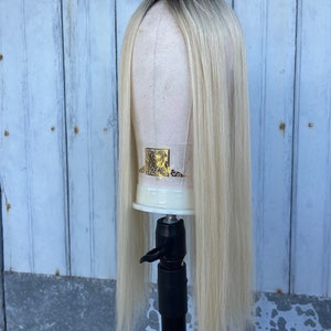 Puede incluir: Peluca rubia con raíces oscuras exhibida en una cabeza de maniquí. La peluca tiene cabello largo y liso que cae por debajo de los hombros. La cabeza del maniquí está montada sobre un soporte negro contra un fondo de madera azul claro desgastado.