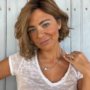 May include: A silver necklace with three circular, gem-studded pendants. The necklace is worn with a white V-neck t-shirt. The person is wearing two silver rings on their right hand.