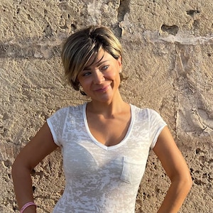May include: A woman wearing a white V-neck t-shirt with a chest pocket. The woman has short, layered blonde and brown hair. She is smiling and standing in front of a textured, light brown wall.