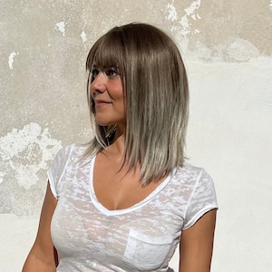 May include: A woman with shoulder-length, ash-blonde hair with bangs. She is wearing a white, semi-sheer V-neck t-shirt with a pocket. The background is a textured, off-white wall.