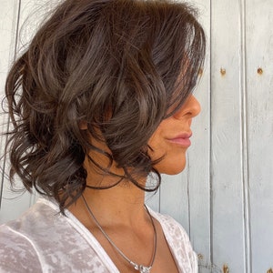 May include: A side profile view of a person with short, wavy, dark brown hair. They are wearing a white, textured top and a silver necklace with a clear, round pendant. The background is a weathered, light-colored wooden wall.