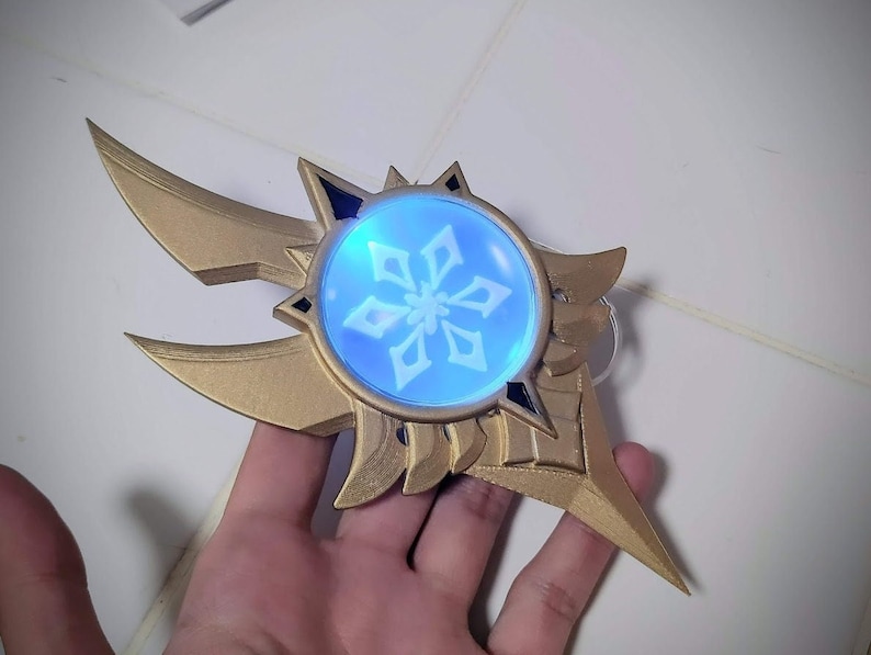 May include: A gold-colored, 3D printed, decorative object with a snowflake design in a blue, translucent circle. The object has a sharp, pointed tip and two wings.