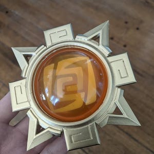 May include: A gold-colored, star-shaped pendant with a circular, orange, translucent center featuring a stylized design. The pendant has a raised, textured surface.