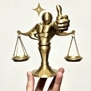 May include: A gold-colored sculpture of a muscular figure holding a scale, with a thumbs-up gesture. The figure has a round head and is standing on a pedestal base. A star-shaped accent is above the figure.