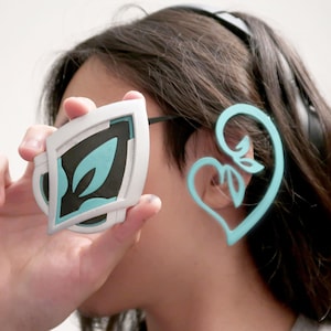 Può includere: Un paio di orecchini a cuffia stampati in 3D di colore blu turchese. Un orecchino ha la forma di un cuore con un design a foglia. L'altro orecchino è di forma geometrica con un design a foglia.