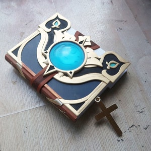 May include: A closed book with a gold and black cover, a turquoise gem, and a gold cross charm attached. The book has a leather strap and ornate details.