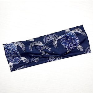 Op de afbeelding: Een marineblauwe hoofdband met een herhaald patroon van witte zeeschildpadden. De schildpadden hebben gedetailleerde schaalontwerpen. De hoofdband heeft een gedraaid knoopontwerp en is gemaakt van een zacht, flexibel materiaal. Kleine witte sterren staan op de achtergrond.