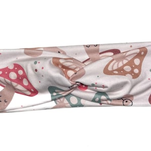 May include: White headband with a repeating pattern of whimsical mushrooms in shades of red, brown, and teal. The headband is twisted at the center and features small, colorful polka dots scattered across the fabric.