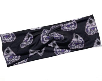 Op de afbeelding: Zwarte hoofdband met een herhalend patroon van paarse planchette- en oogontwerpen. De hoofdband is aan de bovenkant gedraaid en is gemaakt van een zachte, rekbare stof. Het ontwerp heeft gotische en spirituele thema's.