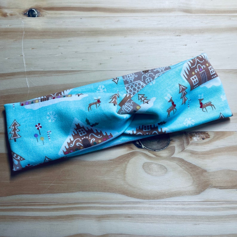 May include: A blue and brown patterned headband with a gingerbread house design. The headband is twisted and has a knot in the center.