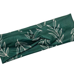 May include: A dark green headband with a twist design, featuring a repeating pattern of white leaf and stem illustrations. The headband is made of a soft fabric and is designed to be worn around the head.