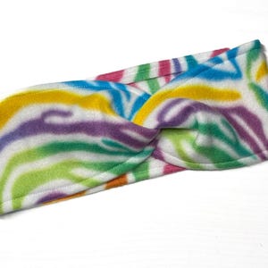 May include: A colorful fleece headband with a twisted knot design. The headband features a vibrant zebra stripe pattern in shades of yellow, purple, green, blue, and pink. The soft fabric is white with the colorful stripes.