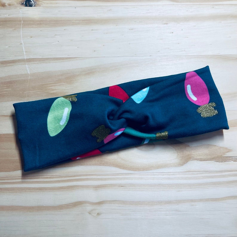 May include: A dark green fabric headband with a Christmas light pattern. The lights are red, green, pink, and blue with gold glitter accents.