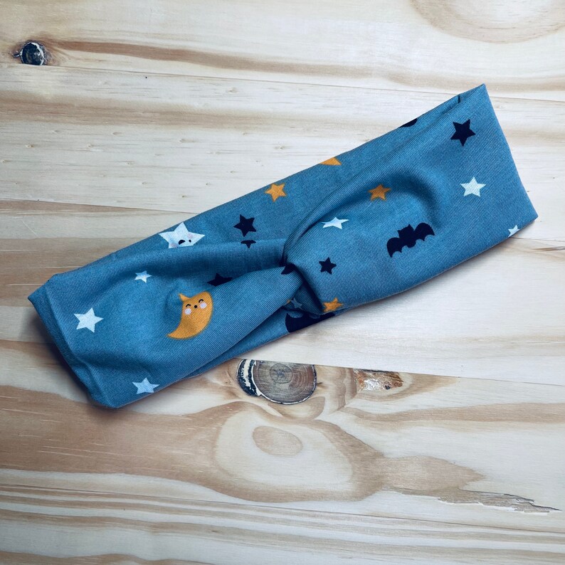 Pode incluir: Uma faixa de cabe&ccedil;a azul com um design de n&oacute; apresenta um padr&atilde;o repetitivo de estrelas, morcegos e luas. A faixa de cabe&ccedil;a &eacute; feita de um tecido macio e el&aacute;stico.