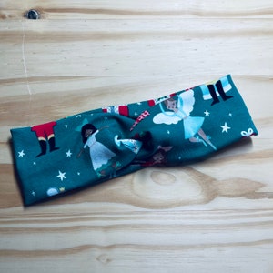 May include: A teal blue fabric headband with a Christmas-themed print featuring a nutcracker, a ballerina, and stars. The headband is twisted and tied in a knot.